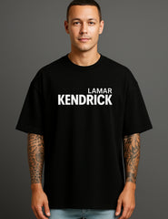 Young man in ATRAYE black streetwear T-shirt with 'Lamar Kendrick' print, tattooed arms.